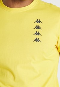 Gelbes Shirt mit vier kleinen schwarzen Logos von zwei Rücken an Rücken sitzenden Figuren, die vertikal auf der linken Seite der Brust gestapelt sind.