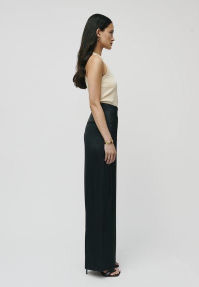 Svarte høyhalsbukser med en glatt tekstur, kombinert med en tettsittende beige tank top. Modellen står i profil mot en ensfarget bakgrunn.