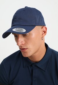 Tumesinine reguleeritav müts koos kumerate visoritega, valmistatud puuvillast. Ees on silt, millel on kiri "The Authentic Adjustable".