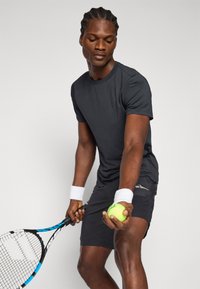 Μαύρο αθλητικό t-shirt και σορτς, κρατώντας μια ρακέτα τένις και μια πράσινη μπάλα τένις, με λευκές χειροστρίφτες που τονίζουν την εμφάνιση.