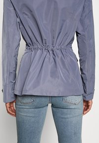 Personne portant une veste violet clair avec taille élastique froncée, vue de dos, associée à un jean en denim bleu clair.