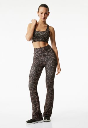 Vrouw staat, gekleed in een bijpassende crop top met luipaardprint en hoge taille legging met zwarte sneakers tegen een witte achtergrond.