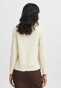 Långärmad, krämfärgad topp i texturerad tyg med ett upphöjt mönster. Designen har en rak fåll och detaljer i sömmen på baksidan.