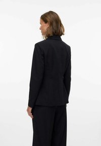 Blazer nero su misura con un design strutturato, rever affilati e tessuto testurizzato, dalla silhouette aderente e chiusura a un bottone.