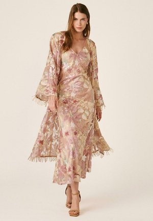 Mujer con vestido rosa con lentejuelas florales, kimono transparente y con flecos a juego y sandalias de tacón alto en color tostado, de pie frente a un fondo neutro.