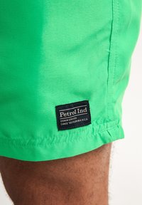Shorts din țesătură verde cu o textură netedă, prezentând o etichetă de marcă neagră "Petrol Ind" pe tiv. Design simplu, fără modele.