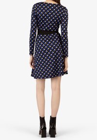 Blaues Kleid mit einem geometrischen Muster aus Dreiecken in Creme und Orange, langen Ärmeln, tailliertem Schnitt mit einem schwarzen Gürtel und einem weit auslaufenden Rock. Schwarze Ankle Boots.