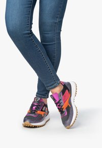 Gambe in jeans blu aderenti con scarpe da ginnastica nere, viola e arancioni con suole bianche, incrociate alle caviglie su uno sfondo bianco.