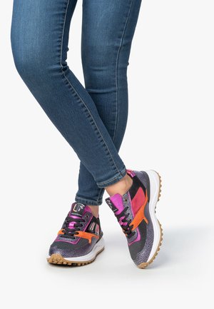 Jambes dans un slim en jeans bleu portant des baskets noires, violettes et oranges avec des semelles blanches, croisées aux chevilles sur un fond blanc.