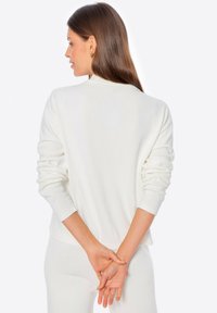 Sudadera blanca con mangas largas y ligeramente abullonadas, y un escote redondo. Textura suave, corte relajado, sin cierres ni patrones visibles.