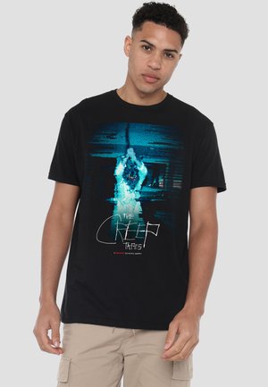 Young man wearing a black T-shirt with a blue glitch-style graphic of a figure hanging and the text "The Creep Tapes."