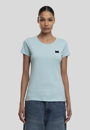 Femme portant un t-shirt bleu clair avec un petit graphique de visage de chat noir sur le côté gauche de la poitrine et un jean bleu sur fond gris.