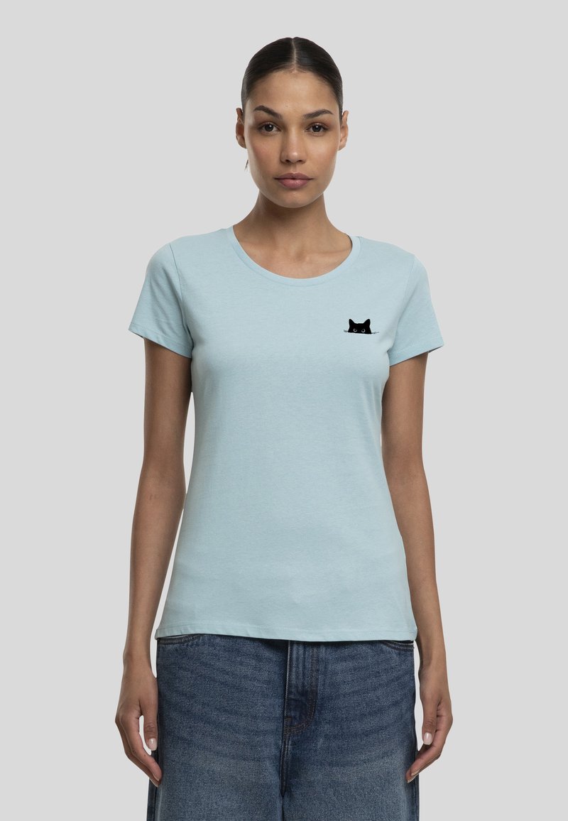 Femme portant un t-shirt bleu clair avec un petit graphique de visage de chat noir sur le côté gauche de la poitrine et un jean bleu sur fond gris.