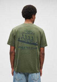 T-shirt de algodão verde-oliva de manga curta com estampa gráfica azul-marinho de quatro personagens de desenho animado num barco com o texto "THE REAL STOCKHOLM ATHLETES CLUB."