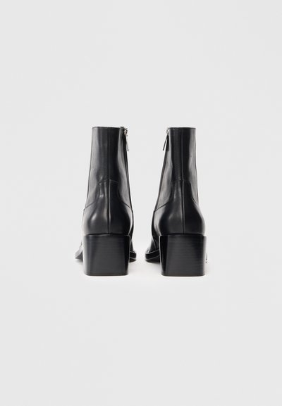 Par crnih kožnih čizama do gležnjeva s blok petama i stražnjim munjevim zatvaračem na bijeloj pozadini.