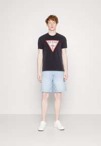 Svart bomulls t-skjorte med rød og hvit trekantet "GUESS" logo, kombinert med lyseblå denimshorts og hvite sneakers.