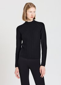 Junge Frau in einem enganliegenden schwarzen Rippenstrickpullover mit langen Ärmeln und schwarzen Hosen, die nach rechts schaut vor einem einfachen weißen Hintergrund.