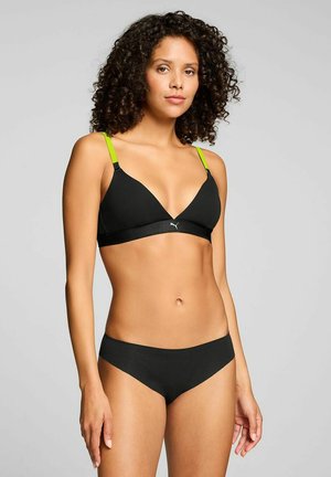 Frau mit lockigem Haar, die einen schwarzen Bikini mit neongrünen Trägern trägt, steht vor einem einfarbigen hellgrauen Hintergrund.