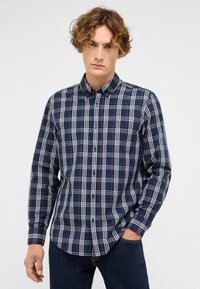 Junger Mann trägt ein kariertes Button-up-Hemd in Marineblau und Weiß mit dunklen Jeans und steht mit einer Hand in der Tasche vor einem schlichten Hintergrund.