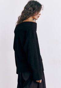 Vrouw met golvend bruin haar, gekleed in een zwarte off-shoulder trui en een zwarte rok, staand in profiel tegen een effen witte achtergrond.
