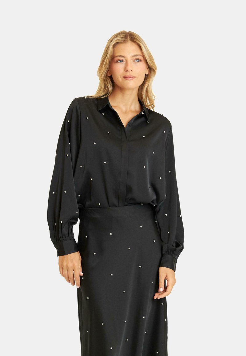 Czarna satynowa bluzka z długimi rękawami, kołnierzykiem oraz zdobieniami przypominającymi perły. Gładka tekstura i luźny krój, zestawiona z pasującą spódnicą.