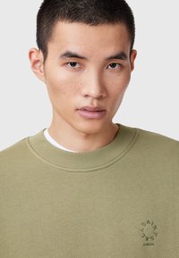 Sudadera verde oliva con cuello redondo, puños acanalados y un logo bordado en el frontal. Fabricada en algodón con textura.