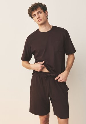 Ung mand iført mørkebrune afslappede shorts og T-shirt, løfter let sin skjorte for at afsløre taljen, neutral baggrund.