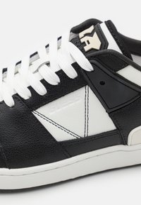 Sneaker bianco e nero in pelle testurizzata, con elementi di design triangolari, lacci bianchi e un accento in plastica nera con logo.