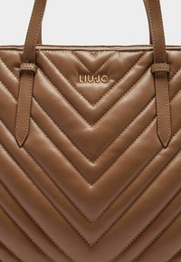 Borsa tote in pelle marrone con trapuntatura a zigzag, con logo dorato e dettagli di cucitura. Le maniglie sono lisce e regolabili.