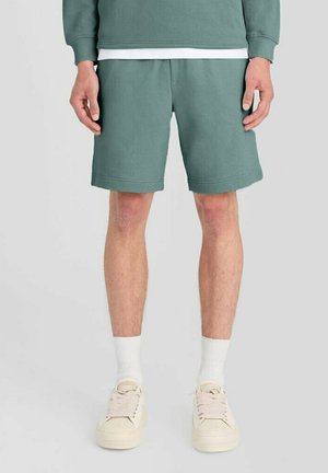 Mand iført grønne shorts, hvide sokker og beige sneakers, der står mod en ensfarvet baggrund.
