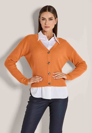 Orange gerippter Cardigan mit V-Ausschnitt, sechs Knöpfen und langen Ärmeln, über einem weißen Hemd mit Kragen getragen und kombiniert mit dunklen Hosen.