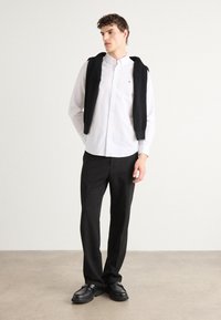 Λευκό πουκάμισο με κουμπιά κάτω από ένα μαύρο αμάνικο πουλόβερ, συνδυασμένο με μαύρες παν trousers και μαύρα loafers. Απλός σχεδιασμός και ουδέτερα χρώματα.