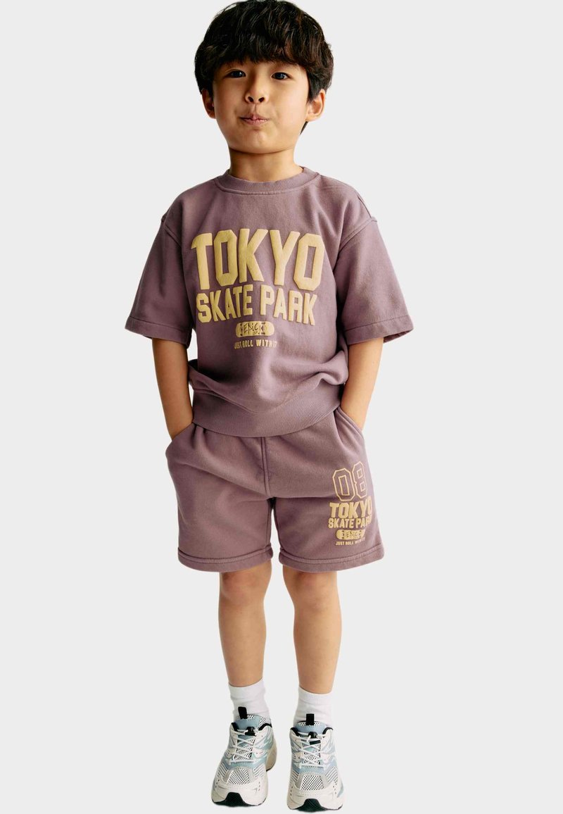 Sudadera de algodón morada de manga corta y pantalones cortos a juego con "TOKYO SKATE PARK" en texto dorado, combinados con zapatillas deportivas blancas y verdes.