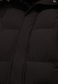 Giacca a vento nera con design trapuntato, caratterizzata da sezioni orizzontali, un colletto alto e una texture liscia. Accentuata con bottoni a pressione.