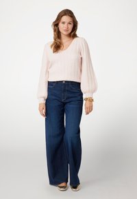 Maglione a coste di un rosa chiaro, con scollo a V e maniche a palloncino, abbinato a jeans a gamba larga di un blu scuro. Accenti dorati sugli accessori.