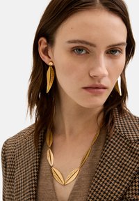 Guldtonet smykker med aflange bladformede designs med ætsede detaljer, parret med en ternet brun blazer og en taupe sweater.