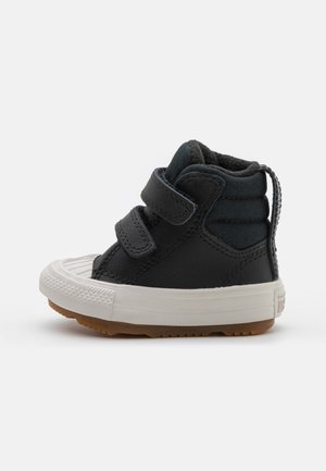 Zapatillas altas negras para bebé, hechas de cuero y lona, con dos correas de velcro, una puntera de goma blanca y una suela de goma color caramelo.