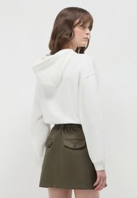 Donna con capelli castani mossi, che indossa un hoodie bianco e una gonna verde oliva con tasche a patta con bottoni, rivolta di lato su uno sfondo semplice.