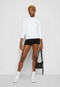 White hoodie with a drawstring, black shorts with a logo, white sneakers, and a small black bag. Smooth fabric, slim fit, minimalist design.