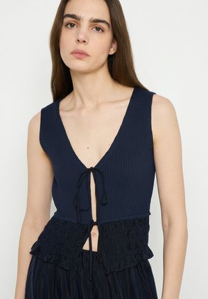 Jeune femme portant un haut en tricot bleu marine sans manches avec un profond col en V et une fermeture devant nouée, associé à une jupe texturée assortie.
