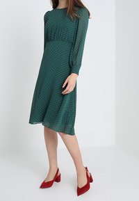 Robe verte à pois avec manches longues et taille cintrée, associée à des chaussures à talons blocs rouges. Tissu lisse et léger.