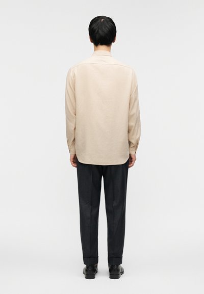 Μπεζ πουκάμισο με μακριά μανίκια και ίσια γραμμή, σε συνδυασμό με σκούρο παντελόνι με λεπτές ρίγες και μαύρα παπούτσια. Το πουκάμισο έχει γιακά με κουμπιά.