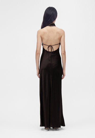 Femme aux longs cheveux noirs porte une robe noire longue jusqu'au sol, dos nu, col halter, nouée avec des fines bretelles à l'arrière, debout sur des talons.