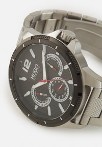 Orologio in acciaio inossidabile nero con quadrante grande, dettagli argentati e particolari rossi. Presenta sottocampi per la data e il giorno. Bracciale con finitura testurizzata.