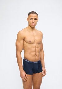 Άνδρας χωρίς μπλούζα με μυώδες σώμα, που φοράει σκούρα μπλε εσώρουχα Calvin Klein, στέκεται μπροστά σε ένα απλό φωτεινό φόντο.