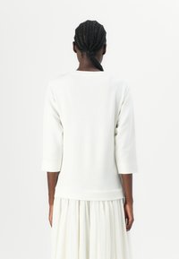 Pullover bianco a maniche lunghe con scollatura rotonda, realizzato in tessuto morbido, con polsini e orlo a coste, indossato sopra una gonna fluida bianca.