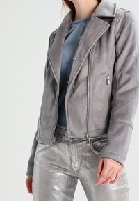 Harmaa mokkanahkainen moottoripyörätakki, jossa on viistetty kaulus, vetoketjulliset taskut ja sovitettu leikkaus, yhdistettynä metallinhohtoisiin hopeisiin housuihin.