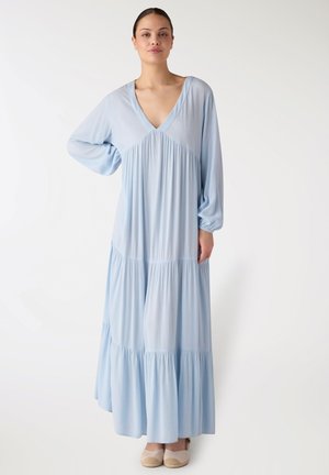 Donna in piedi che indossa un lungo vestito azzurro chiaro a balze con maniche lunghe e scarpe espadrillas beige.