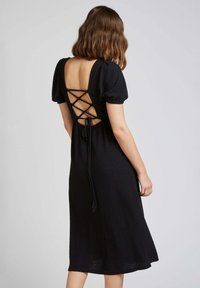 Robe noire en tissu texturé, avec des manches ballon et un dos lacé. La robe présente un corsage ajusté et une jupe fluide.
