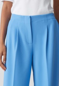 Lichtblauwe broek van een gladde stof, met een voorplooi, tailleband en zijzakken, gezien boven een witte top.